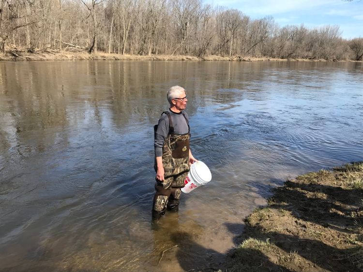 St. John's Lutheran School teacher Mark Koschmann stands in a river wearing waders and holding a pail in his hand.