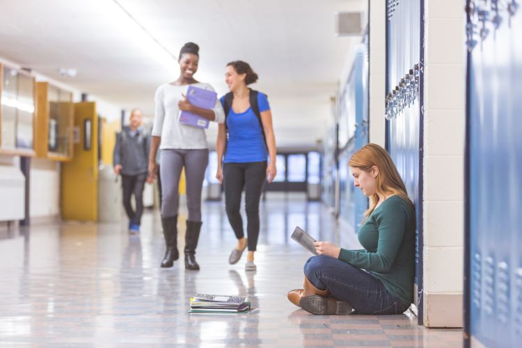 A female student sits in the hallway with her back to the lockers and studies while other students walk past her.