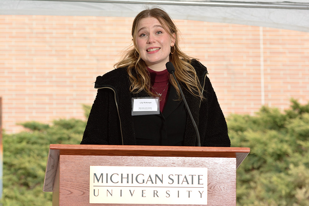 Lily Kirkman speaking at a podium with the Michigan State University wordmark on the front.