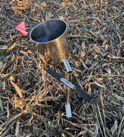 A soil probe imbedded in a field of corn stubble with a stainless steel bucket containing a pink flag.