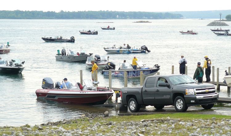 A number of fishing boats are seen on a river. A pickup truck is backed up to the lake at a boat launch and launching a boat .