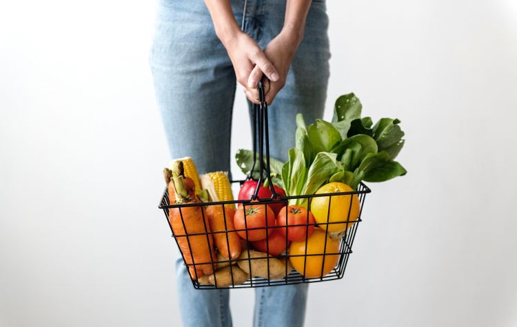 A grocery basket filled with carrots, corn, oranges, tomatoes and leafy greens.