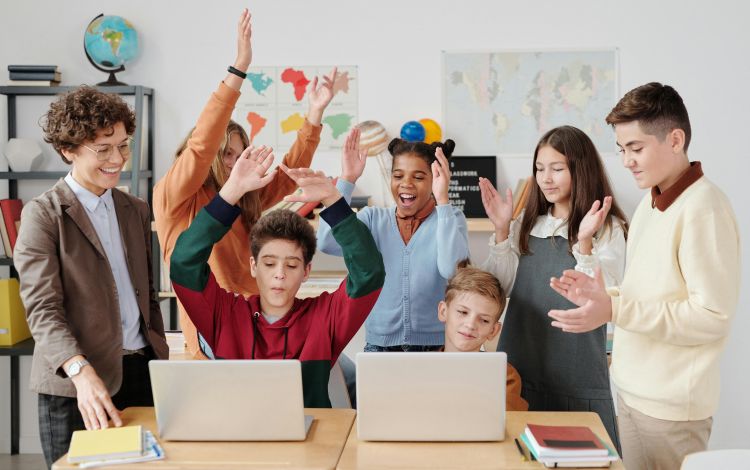 Two youth at laptops with a group of cheering teens and a teacher next to them.
