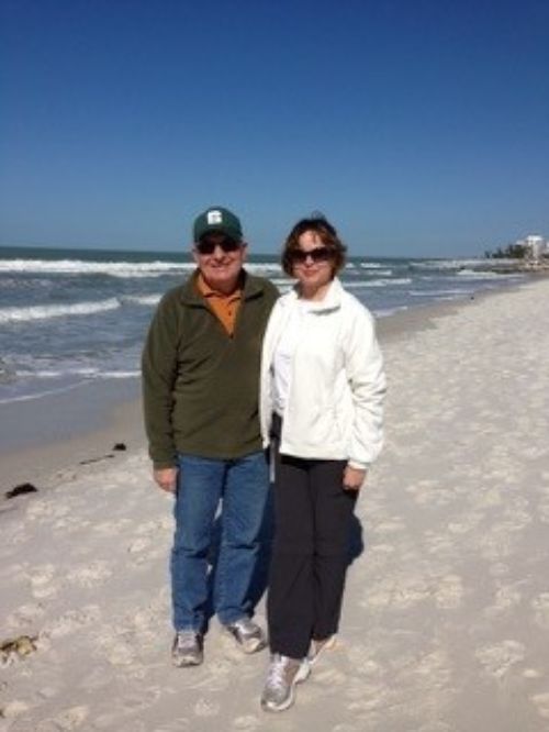 A many in a green shirt and MSU hat and a woman in a white jacket and sunglasses on the beach with water in the background.