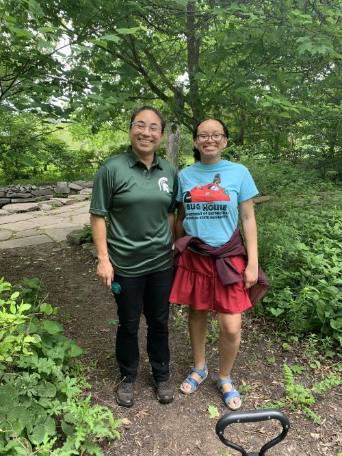 Two woman stand next to each other, smiling at the camera, in a wooded area.