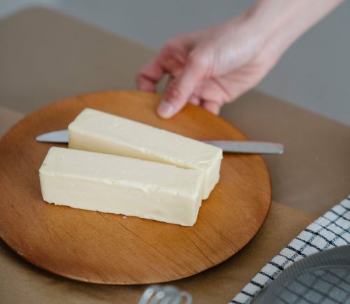 A photo of two sticks of butter with a knife on a small wooden plate being placed on a table by a hand.
