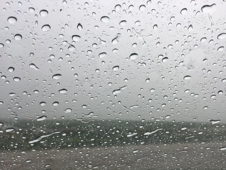 Close-up view of raindrops covering a window, with a blurred agricultural field and gray overcast sky visible in the background.