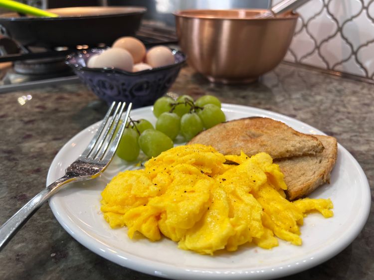 A photo of scrambled eggs on a plate next to toast and grapes.