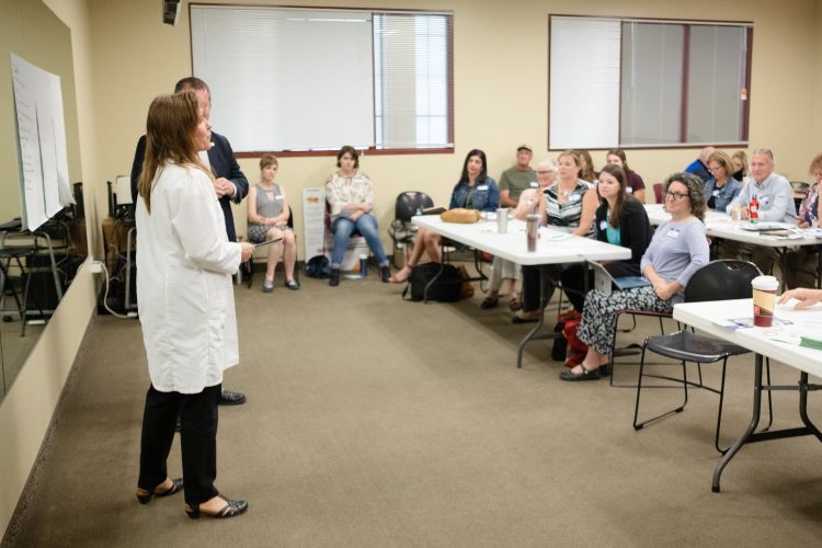 A group of people sit at tables to listen to two people speak to the whole group.