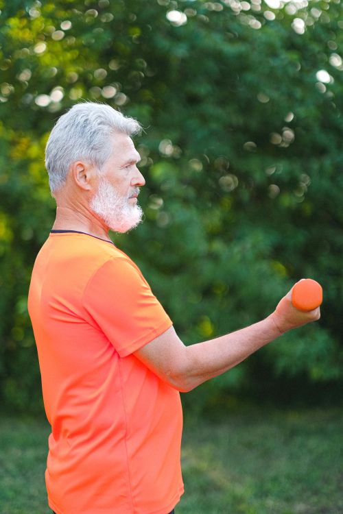 An older adult in an orange shirt practices lifting a light weight outside.