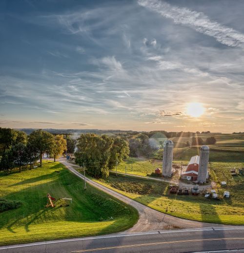 A scenic scene of a farm with a barn and silos and the sun setting behind it.