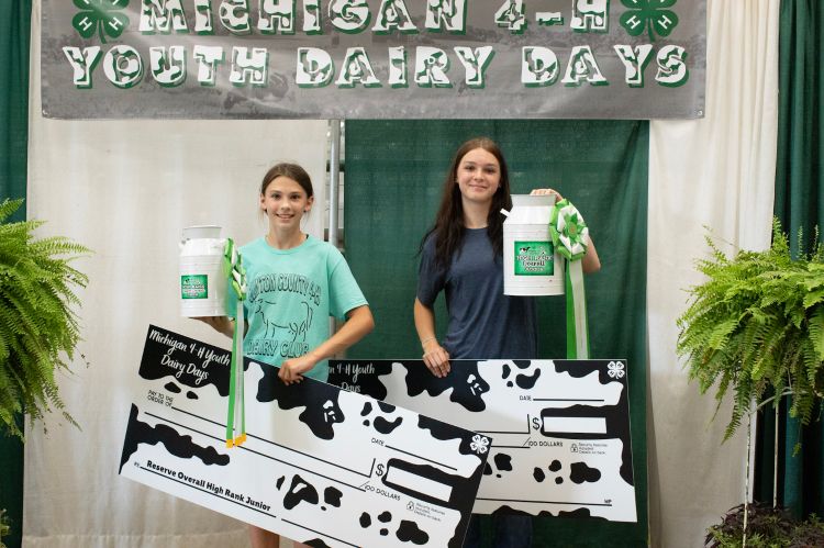 Two youth holding up black and white checks and prizes in front of a Michigan 4-H Youth Dairy Days banner.