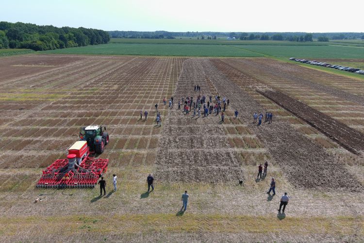 red tiller behind green tractor in brown field with people watching