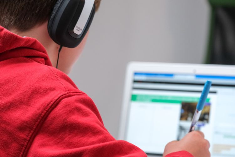 Individual writing notes with a pen with over-the-ear headphones on and looking at a computer screen.