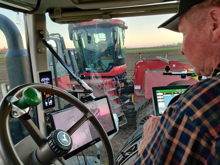 A farmer sits in the cab of a tractor equipped with multiple digital displays and control screens, monitoring precision agriculture equipment. Through the windshield, another large red farming machine is visible working in a field at sunset.