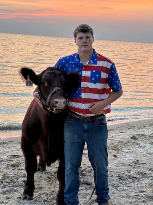 A young man in a red white and blue shirt and jeans standing on the beach with the lake behind him. He is holding a red steer.