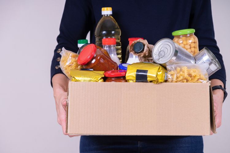 A person holding a box full of pantry items, including canned food and pasta.