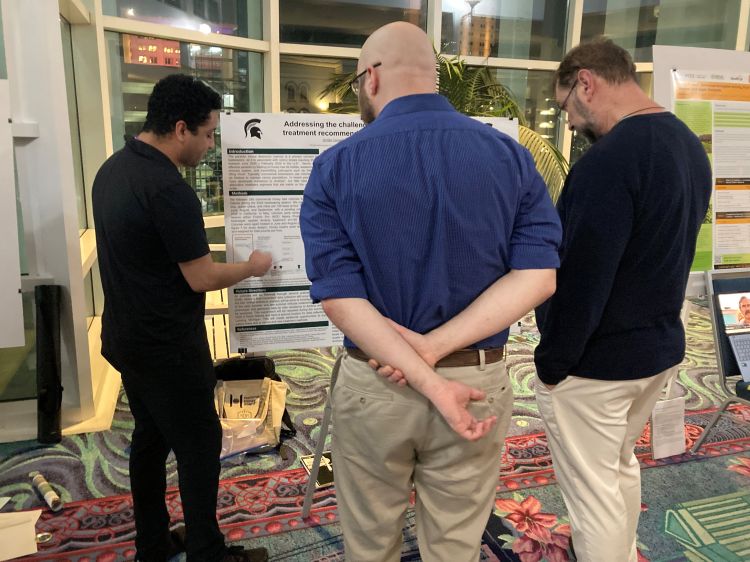 Two people standing in front of a detailed poster while a third person is standing next to the poster pointing at it and talking.