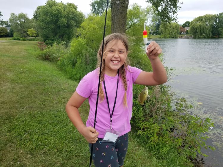 A young girl in a pink shirt smiles while standing at the side of a river holding a fishing pole in her right hand and holding up a fish still on the line.