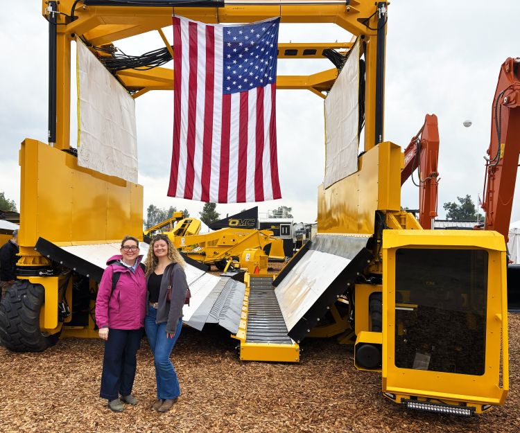 Two women pose for a picture in front a large piece of orchard equipment, with an American flag hanging from the top.