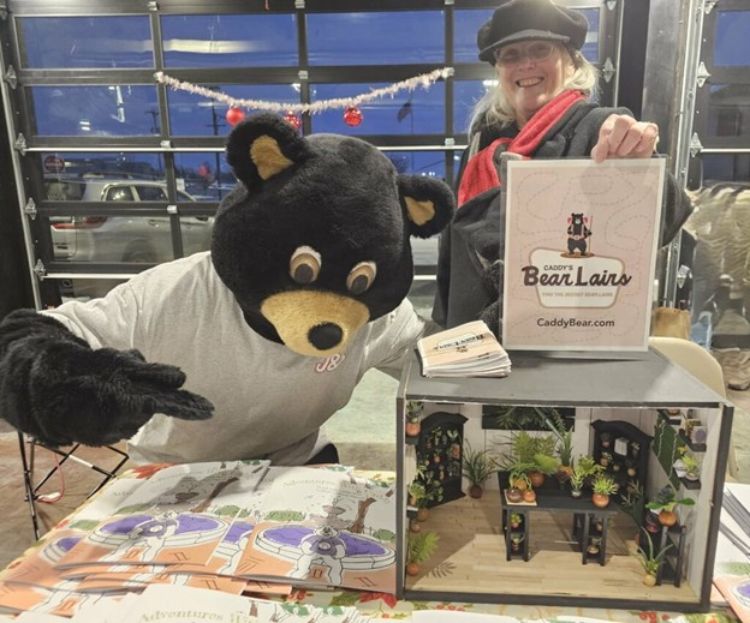 A bear mascot pointing to a diorama. Next to the diorama stands a woman holding up a Bear's Lairs sign.