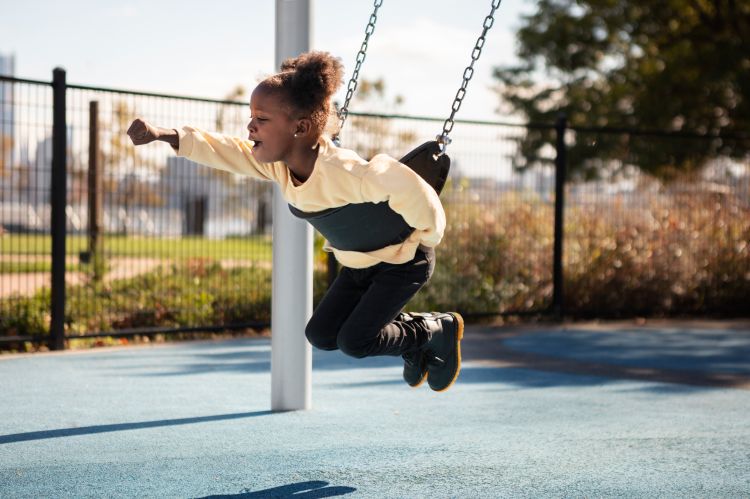 A child swings belly first on a swing at the playground and holds out her arm. The child appears excited and happy.