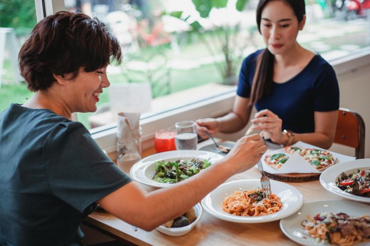 Two people sit across from each other while eating at a restaurant, the food includes salad, pasta, and pizza.