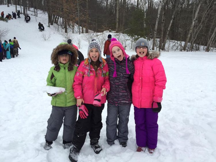 A group of young girls in colorful snow gear standing in the snow with the woods behind them.