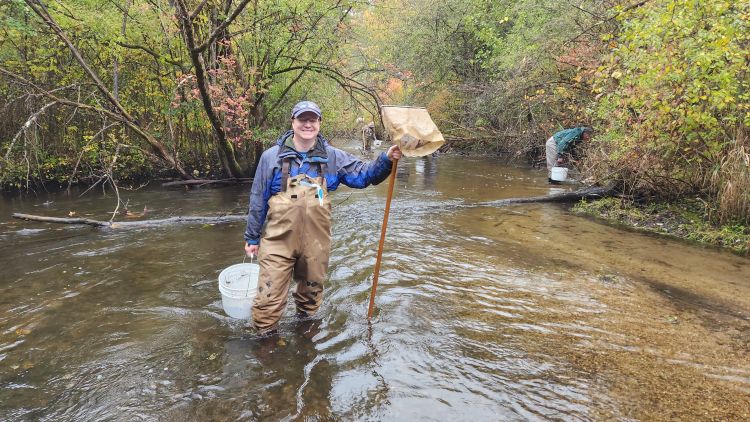 A smiling MiCorps volunteer stands in a stream in waders, carrying a net and bucket.