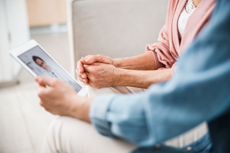 Two people, with only arms and chests visible, look down at a tablet screen showing a doctor delivering telehealth services.