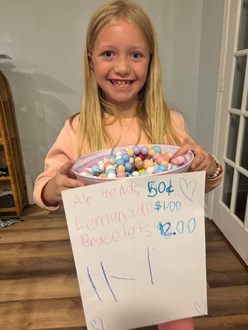 A little blonde girl in a pink shirt with a sign with prices on it.