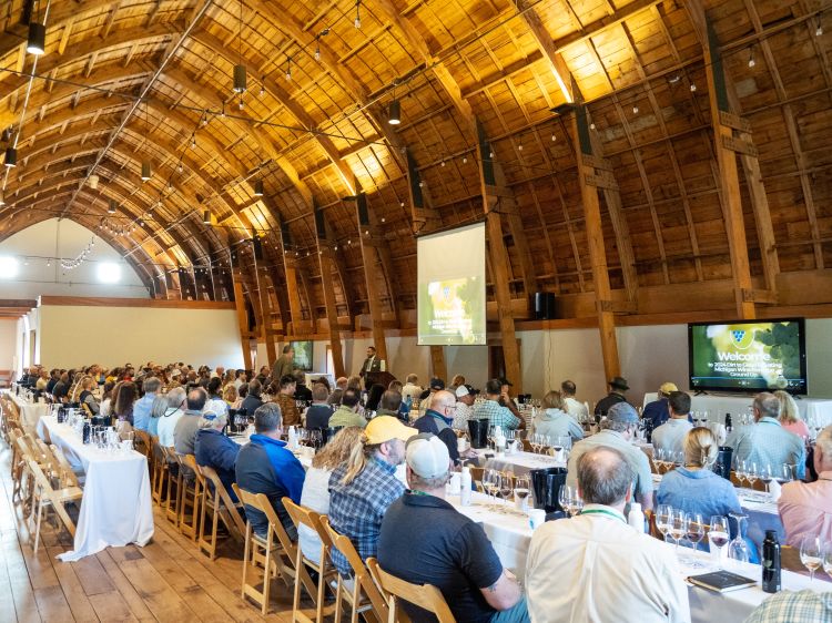 A man stands in front of a room inside of a historic barn, delivering the opening remarks to lots of people sitting at long tables with white table cloths.