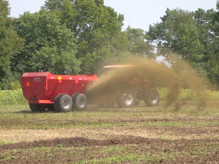 Red tractor pulling red manure spreader that is casting manure on a green and brown field.