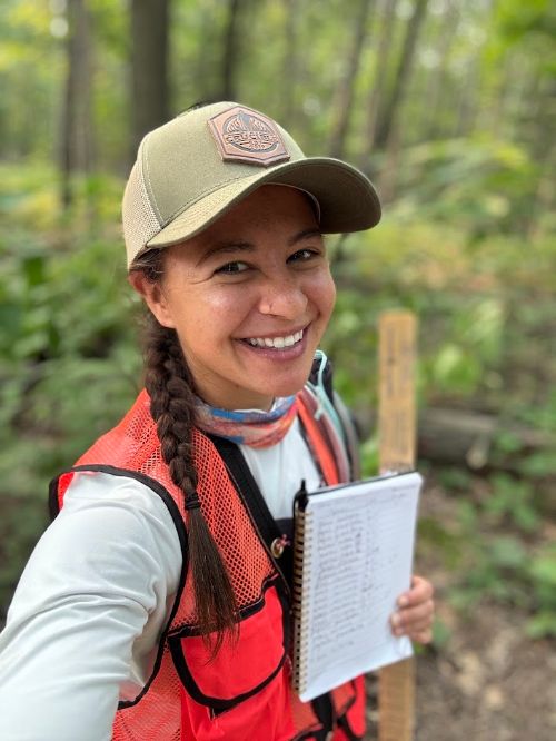 A woman wearing a hat and red vest, posing for a selfie with a forest in the background.