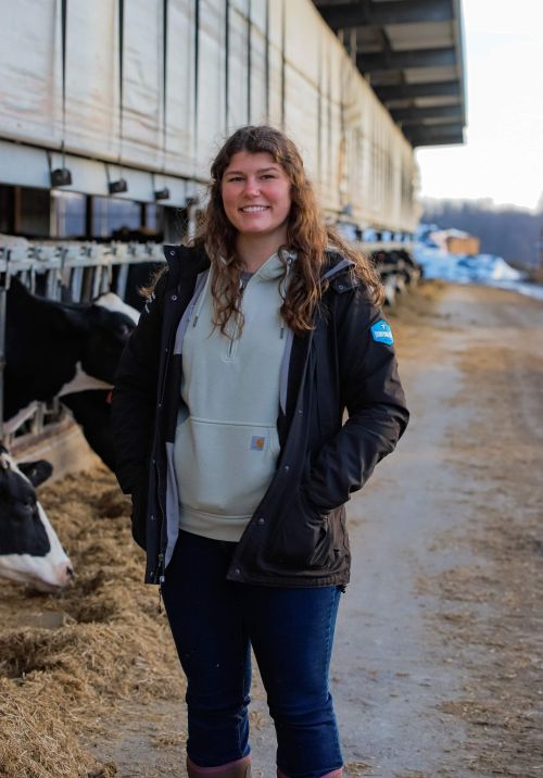 A smiling woman stands in a dairy barn aisle wearing a dark jacket over a light green hoodie and jeans, with cows feeding behind her along the left side and a sunlit farmyard visible in the background.