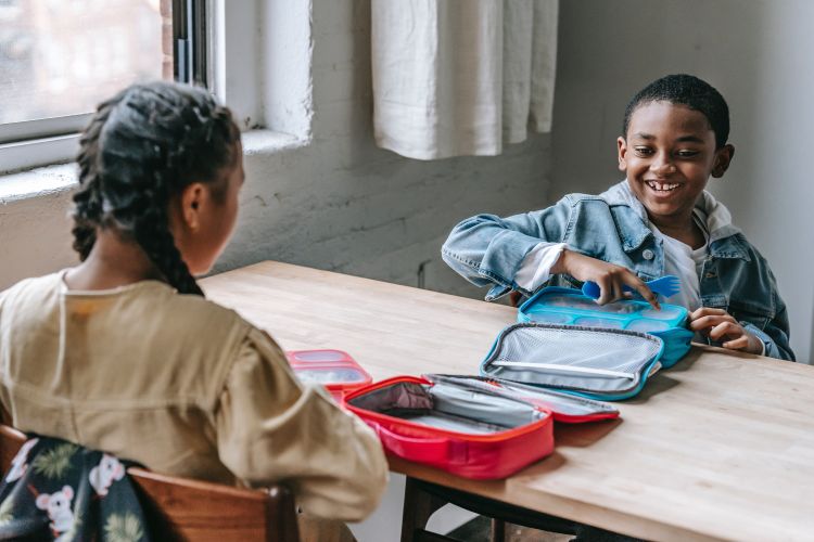 Two happy school-aged children eating lunch together out of lunch boxes.