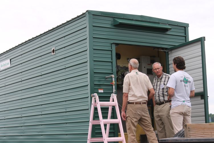 Example of a firewood kiln. Long green structure with metal siding, three individuals standing in front of the door in the front of the structure.