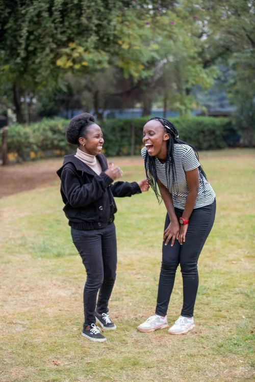 A photo of two women laughing together while standing outside on green grass.
