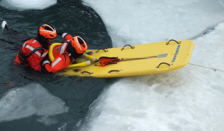 Two members of the Coast Guard are in the water demonstrating the rescue technique of how one person places another, who has apparently fallen through the ice, on a yellow rescue sled to get them out of the water. Photo credit: Friends of the Detroit River.