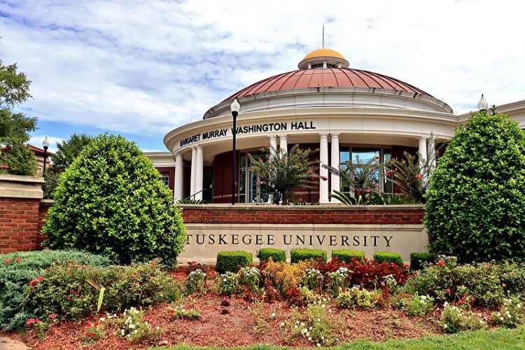 Photograph of Margaret Murray Washington Hall at Tuskegee University, showcasing a circular building with white columns and a red dome roof. The foreground features a landscaped garden with various green shrubs and colorful flowers, with clear signage of the university name on a brick wall.