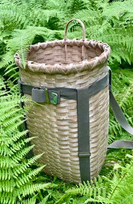 Traditional woven ash basket pack with shoulder straps placed among green ferns outdoors.