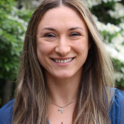 Outdoor portrait of Olivia Spagnuolo standing in front of green foliage with white flowers in the background.