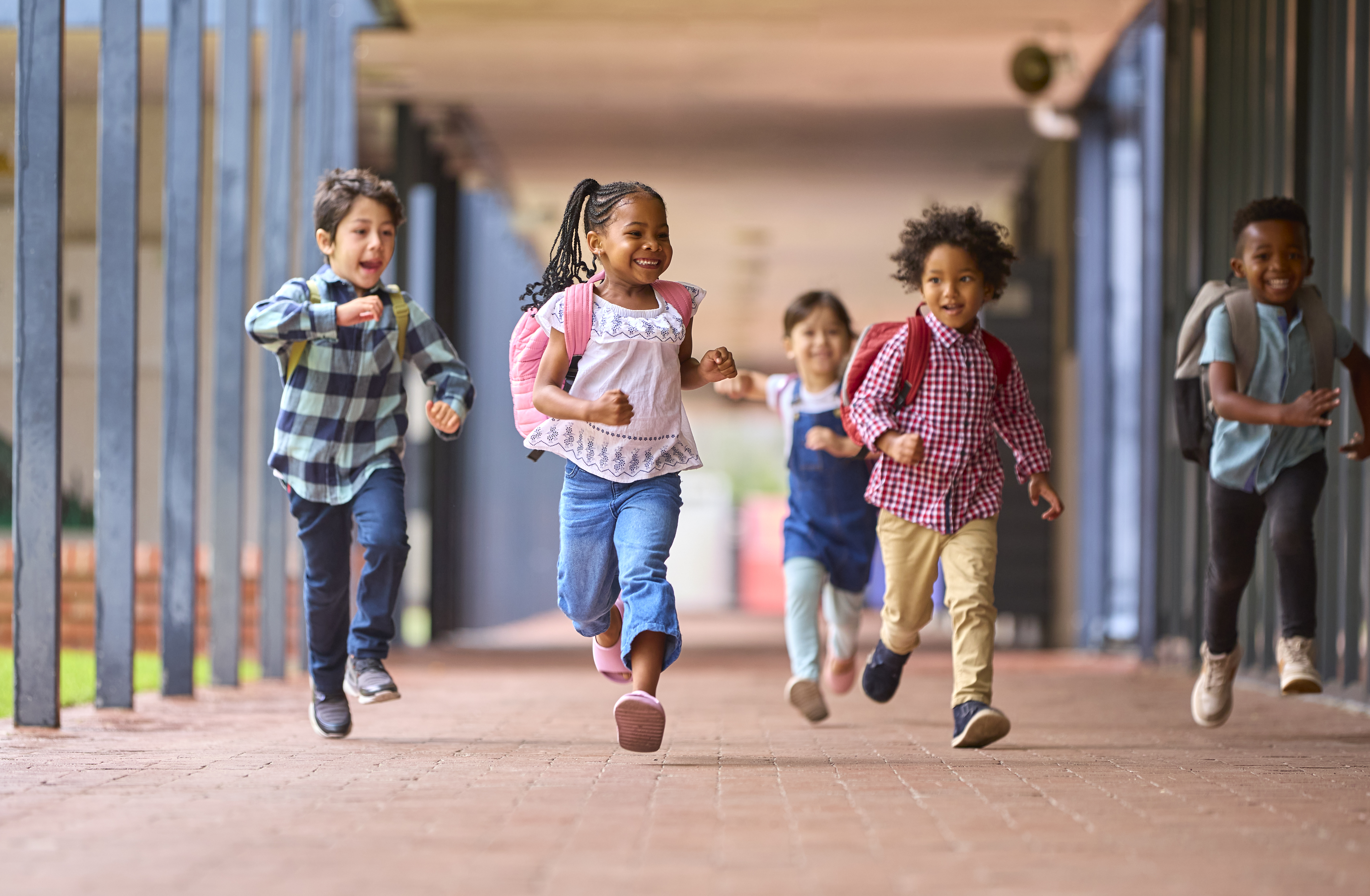 Five young children with backpacks are running down a covered outdoor school walkway, smiling and appearing excited.