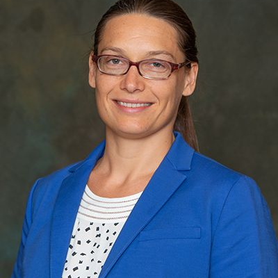 Studio portrait of Hanne Hoffmann wearing glasses, a blue blazer and a patterned white shirt, facing the camera against a neutral dark background.