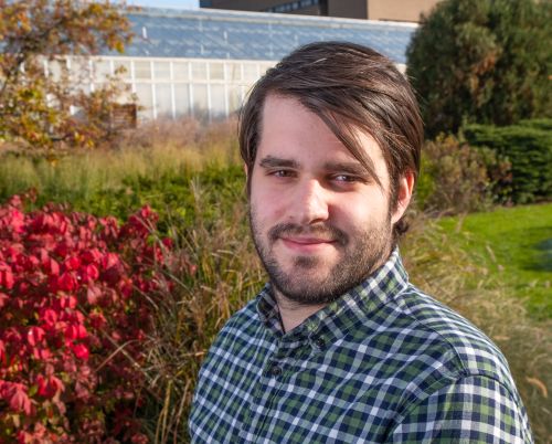 Outdoor portrait of Robert VanBuren wearing a green plaid shirt, standing in front of plants and a building in the background.